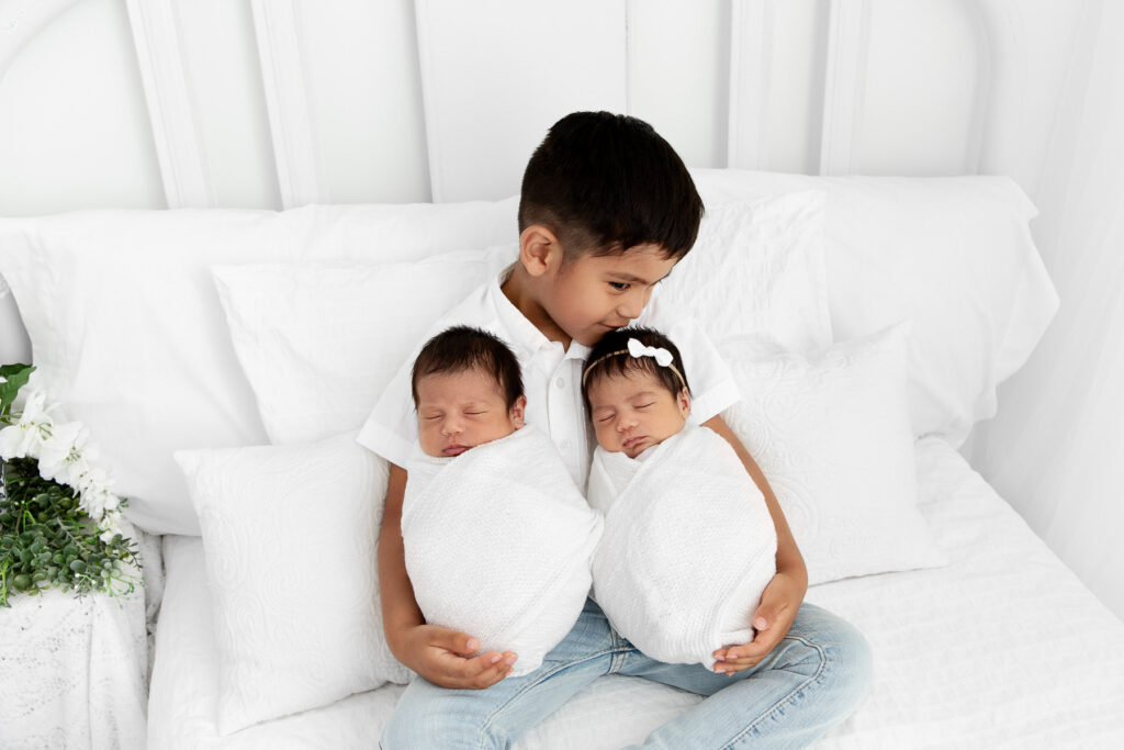 big brother holding swaddled newborn siblings on a white lifestyle bed while kissing little sister's head