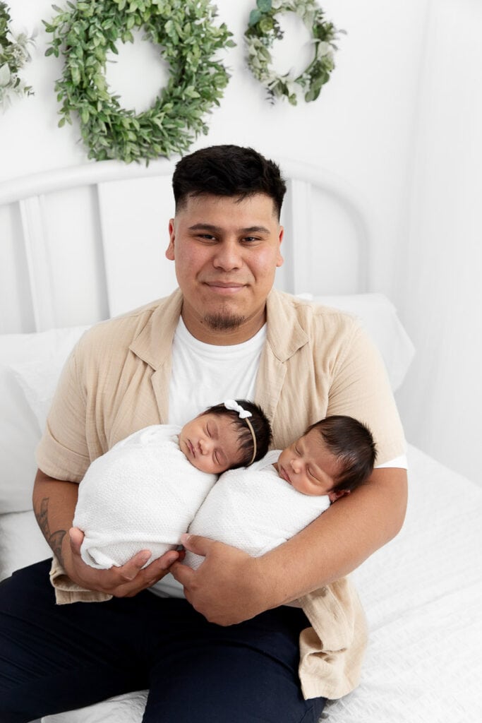 dad sitting on a white lifestyle bed holding swaddled newborn baby twins