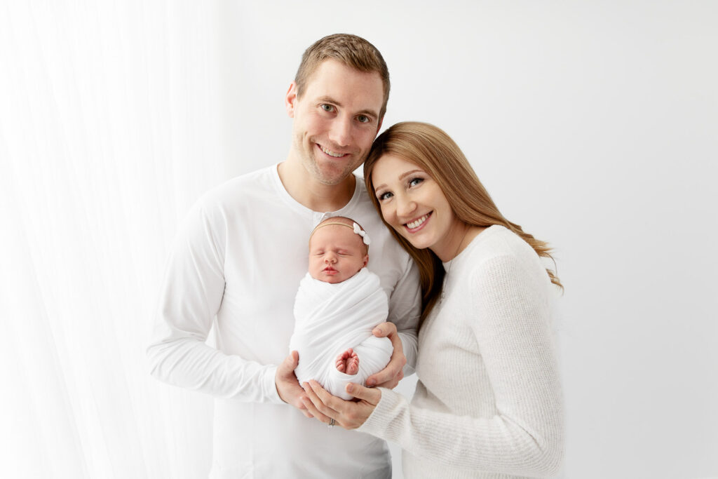 Mom and dad holding swaddled newborn girl in front of sheer white curtain