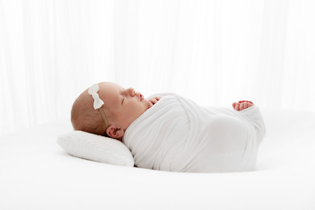 backlit image of newborn baby girl sleeping on a white pillow with toes peeking out of the swaddle