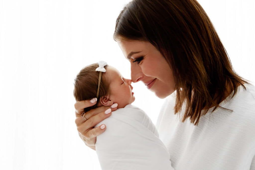 backlit mom holding baby girl nose to nose in front of white curtain