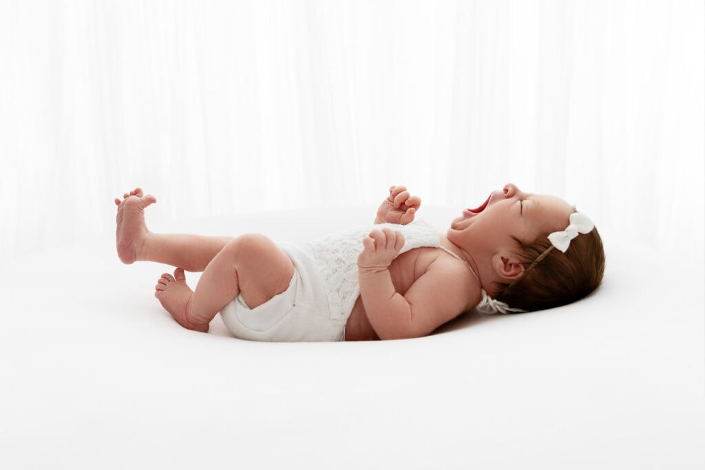 backlit image of newborn baby girl stretched out and yawning in front of white sheer curtain