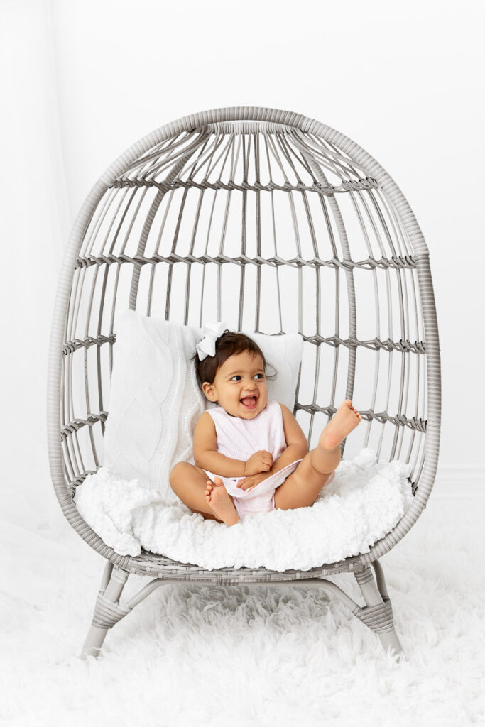 1 year old girl sitting in an egg chair prop laughing with feet in the air