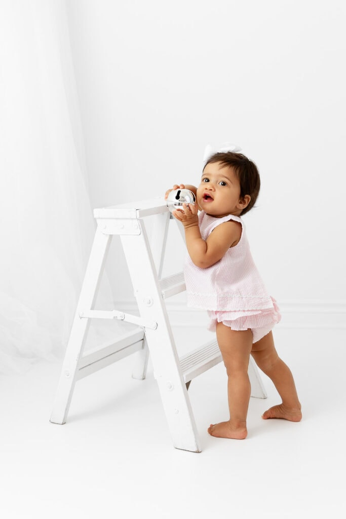 1 year old girl playing with silver bell standing against a white wood ladder