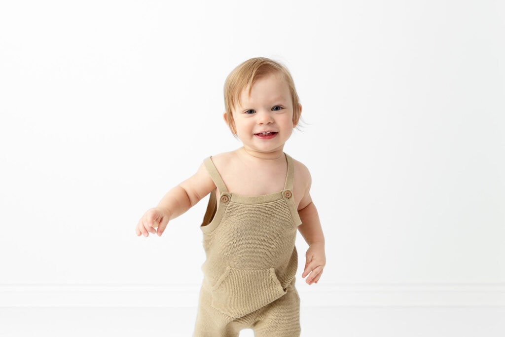 1 year old boy wearing green knit overalls smiling and running towards camera