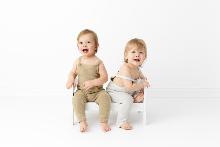 twin 1 year old boys wearing knit overalls sitting together on a white wood doll bed