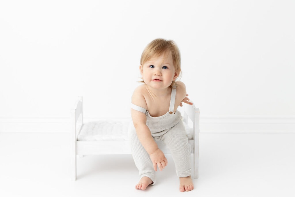 1 year old boy wearing gray knit overalls sitting on a white wood doll bed