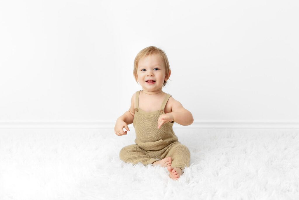 1 year boy wearing green knit overalls sitting happily on a white flokati fur