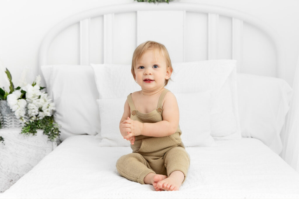 1 year boy wearing green knit overalls sitting on a white lifestyle bed