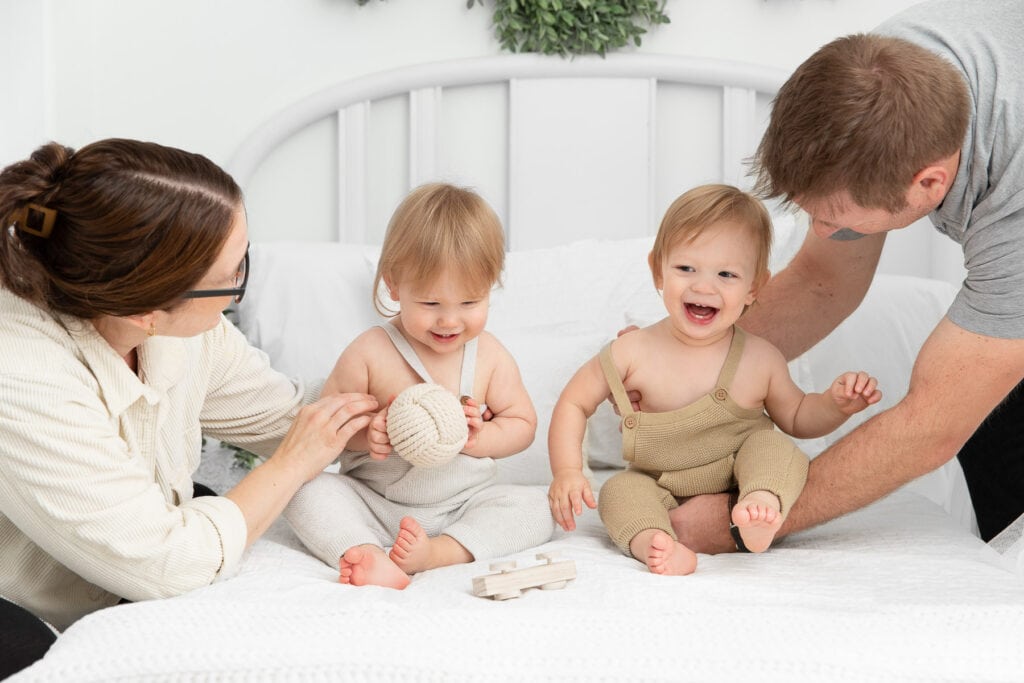 mom and dad with twin 1 year old boys playing on a white lifestyle bed