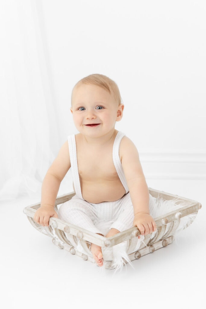 baby boy wearing white overalls sitting in a neutral wood basket