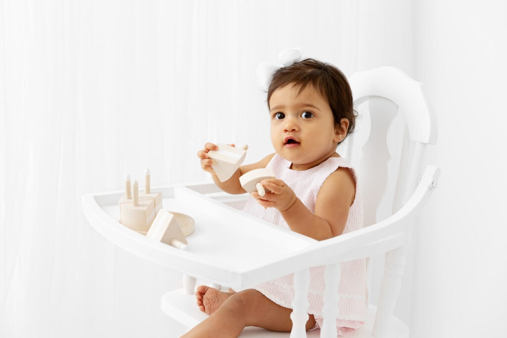 1 year old girl sitting in a vintage high chair playing with a natural wood cake prop