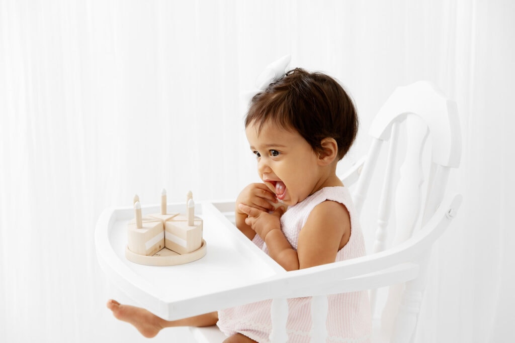 backlit image of 1 year old girl sitting in a vintage white high chair with a wood cake prop
