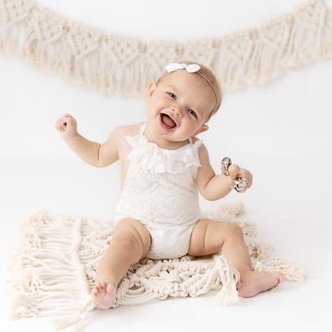 6 month old baby girl smiling and dancing for the camera while sitting on a macrame rug wearing a white romper