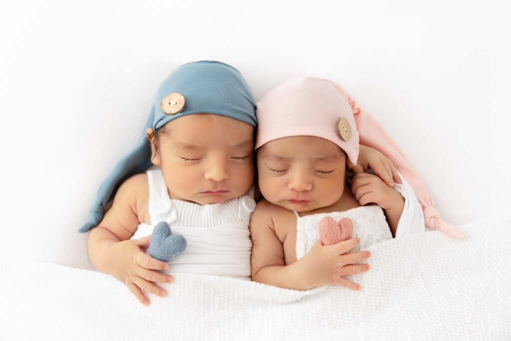 newborn twins wearing blue and pink sleeping caps snuggled together on white beanbag