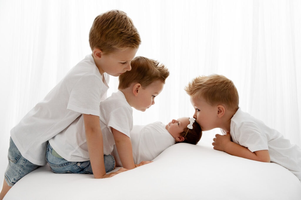 3 brothers looking down at newborn baby sister backlit in front of white curtain