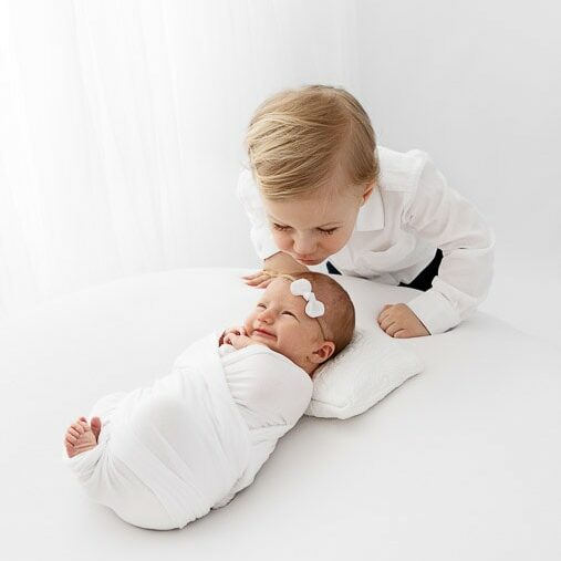 big brother kissing the forehead of smiling newborn baby sister on a white beanbag in front of a white curtain