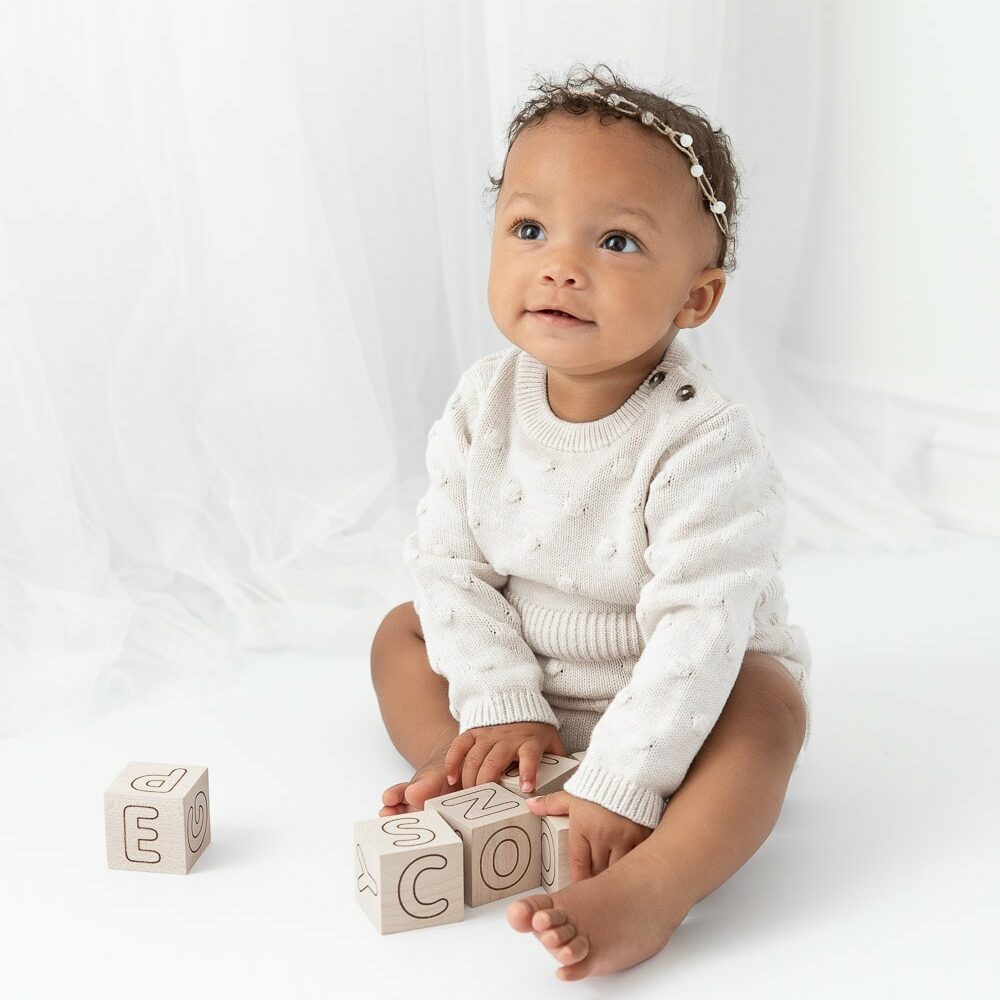 9 month old baby girl sitting in front of a sheer white curtain wearing a white knit romper playing with wooden blocks