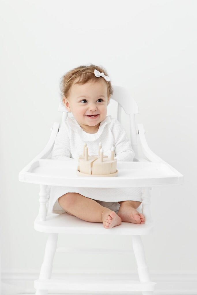 1 year baby girl sitting in vintage white high chair playing with wooden cake