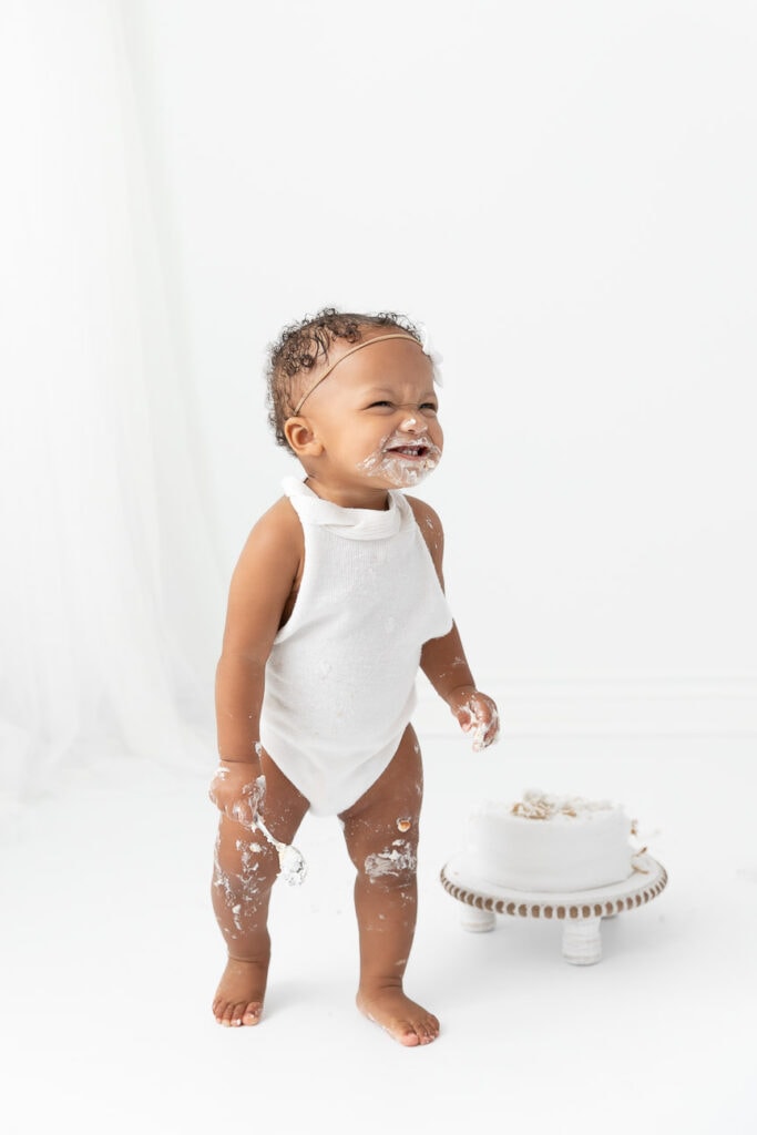 1 year old girl standing near smashed cake coveerd in frosting