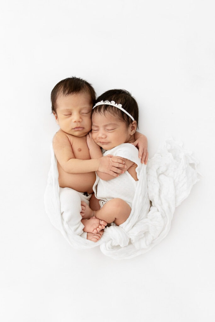 newborn twins snuggled together smiling on white fabric