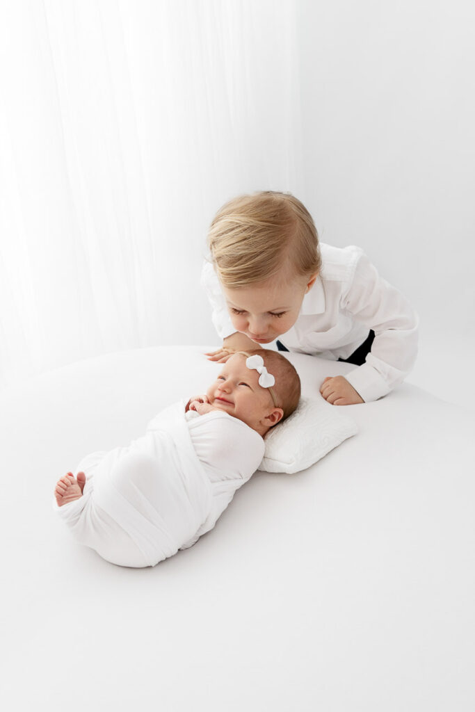 toddler brother kissing newborn baby sister in front of white sheer curtain
