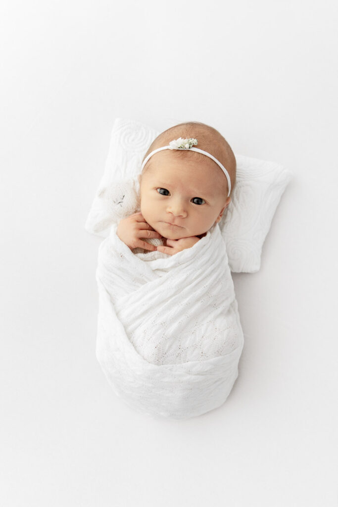 newborn baby girl swaddled and holding a knit bear staring up at camera on a white pillow