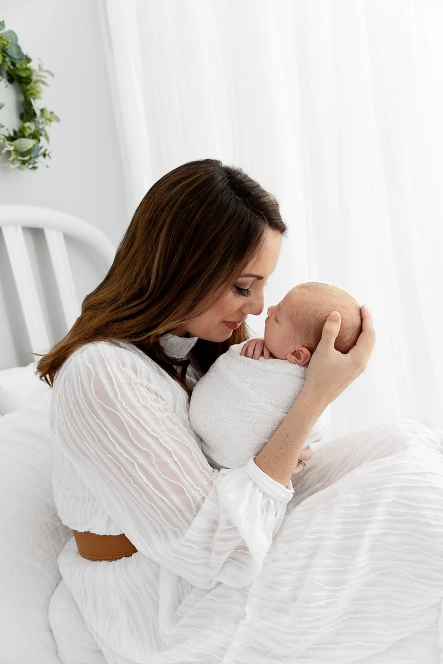 mom holding newborn son close on a lifestyle bed in front of a white curtain